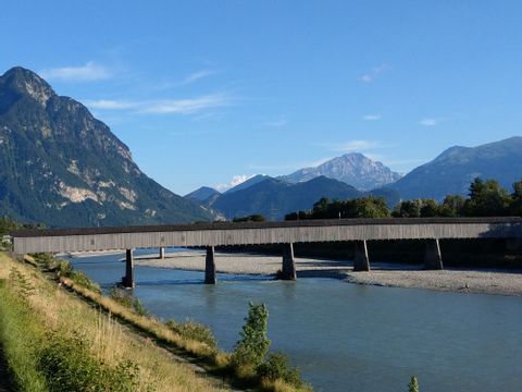 A wooden bridge crosses a river with mountains in the background. 