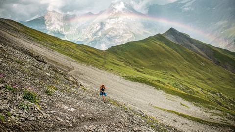 A hiker stands on the Alpschelegrat ridge above Kandersteg. A rainbow shines over the mountain peaks.