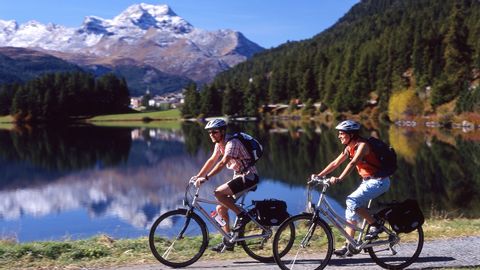 2 bikers ride along the shore of Lake Silvaplana. In the background the snow-covered mountain peaks, which are also reflected in the lake.